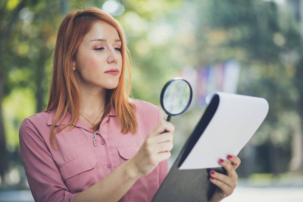 Red-headed woman in a pink blouse holding magnifying glass to read.